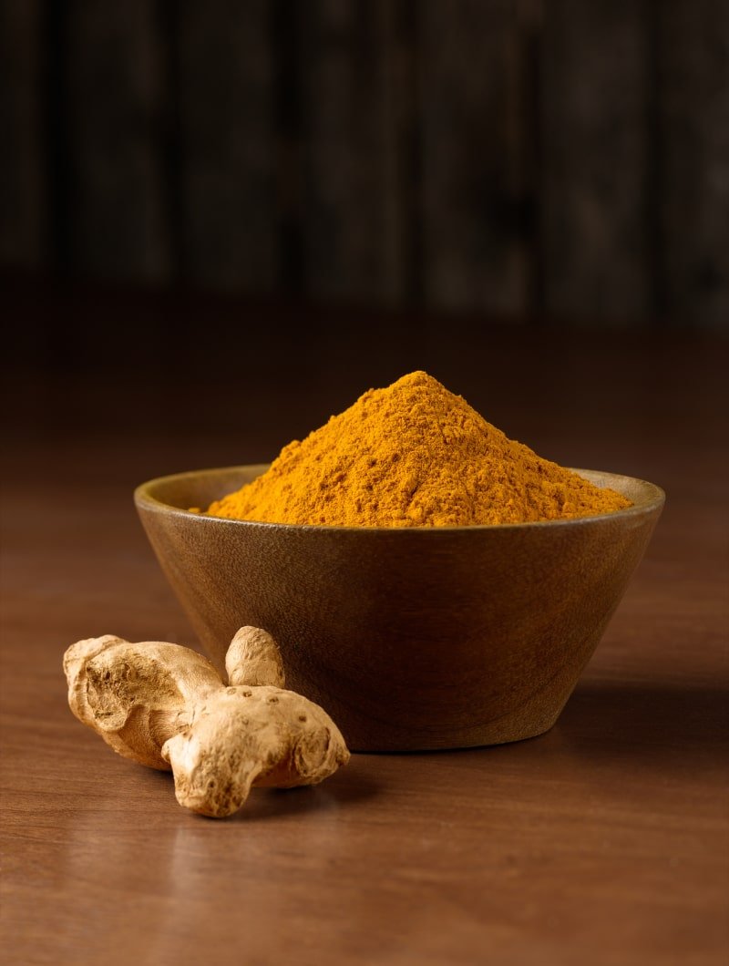 Wooden bowl filled with turmeric powder next to dried turmeric root