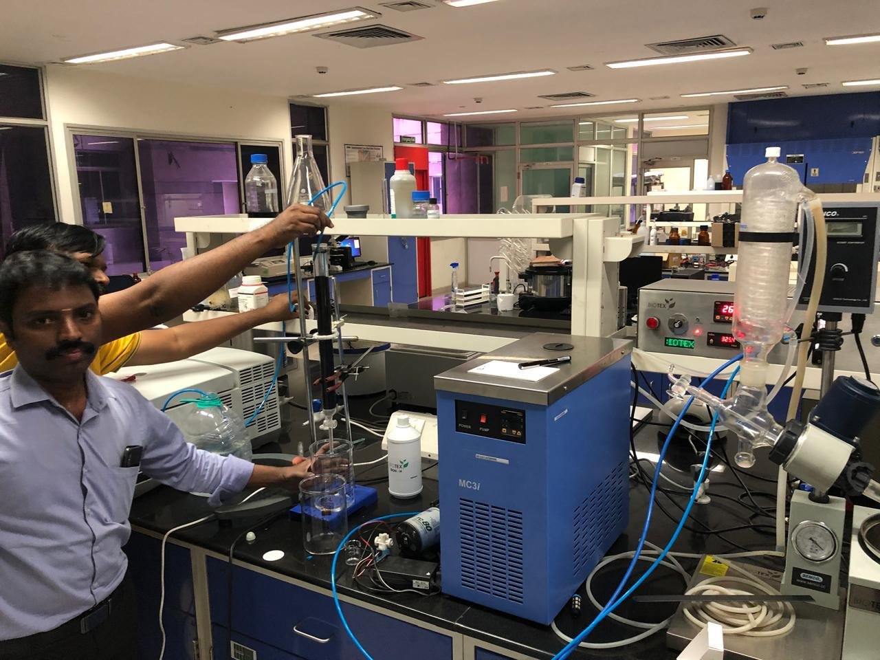 Two male lab technicians working at a laboratory bench with liquid processing equipment including a blue MC3i chiller