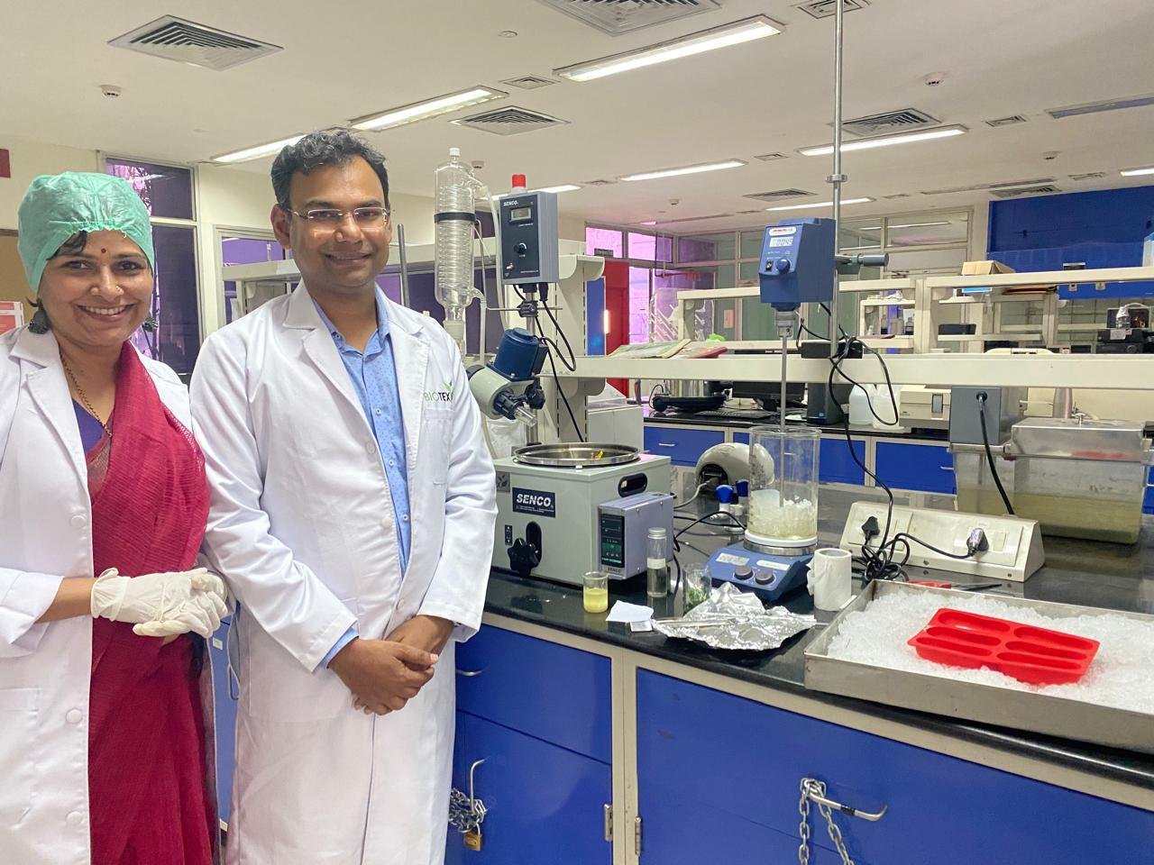Male and female scientists smiling next to laboratory equipment, with the male wearing a white lab coat