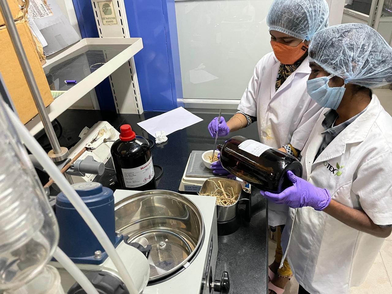 Two female lab researchers wearing hairnets, masks, and gloves working together with a mortar, pestle, and amber glass bottle