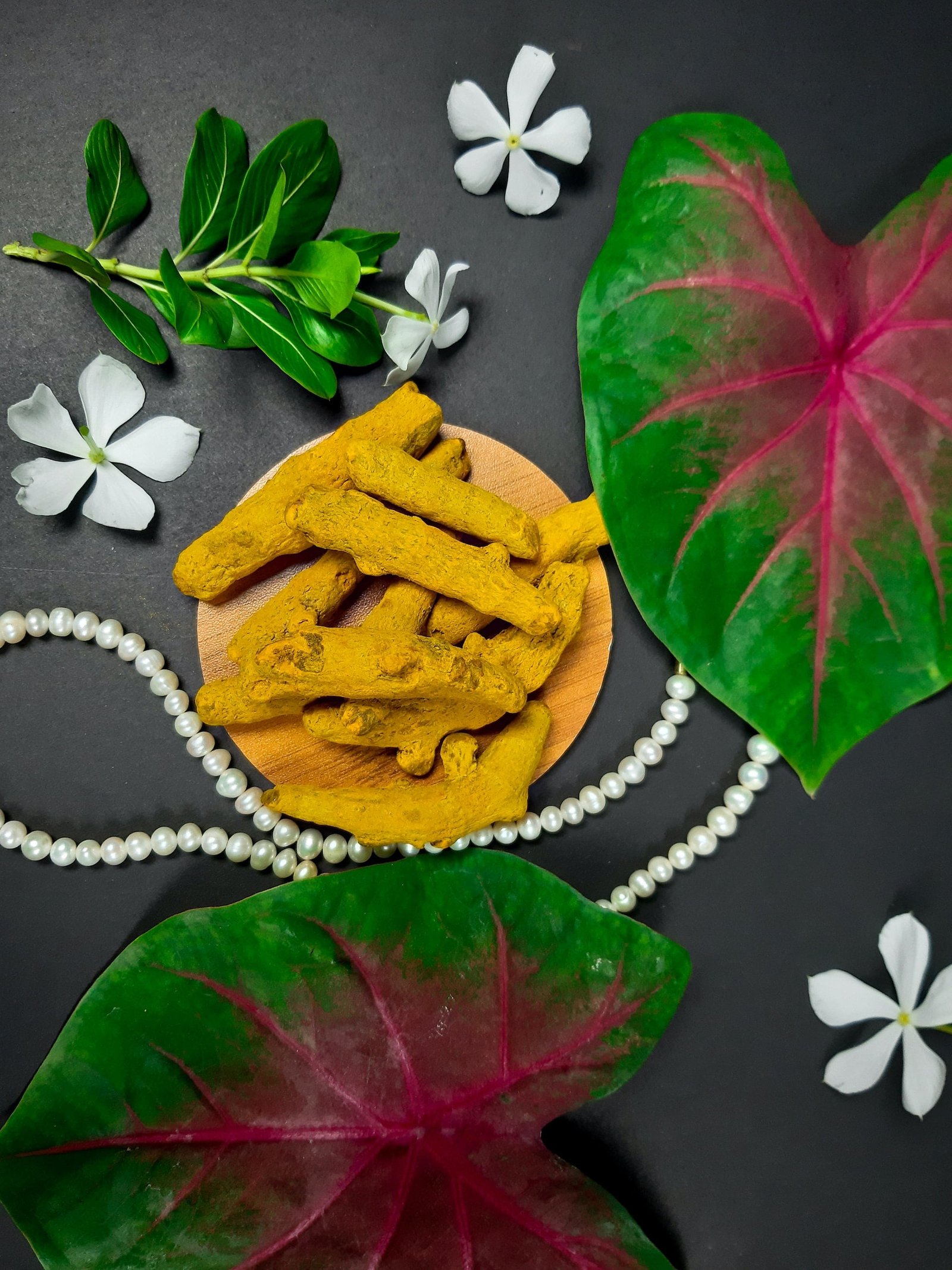 Turmeric roots on a wooden plate surrounded by leaves, flowers, and pearls